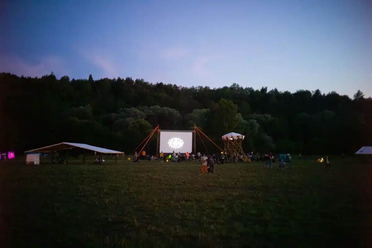 Outdoor amphitheatre filled with audience members watching a film projection on a large screen with orchestral musicians