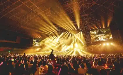 Multidão de fãs em estádio durante show ao anoitecer, com mãos erguidas em direção ao palco